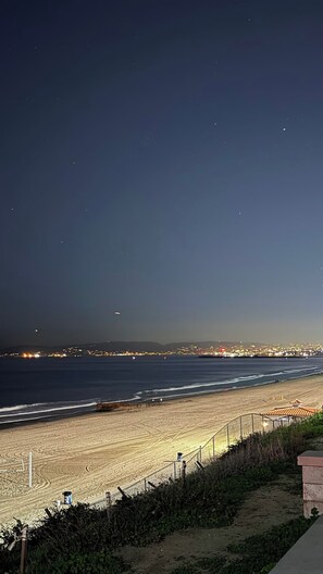 Une plage la nuit, avec la silhouette d’une ville qui se dessine à l’horizon, un sentier sablonneux menant à la plage et une clôture qui longe le bord.