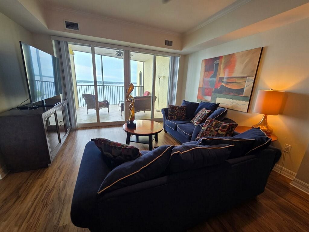 A living room with a sofa, a coffee table, and a TV. There is a balcony with a chair and a table.