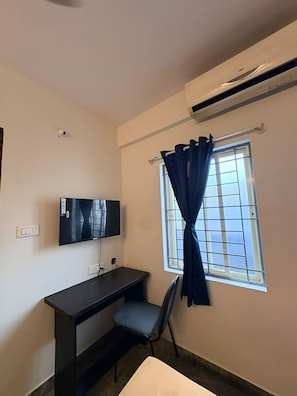 Una habitación con escritorio, silla, televisión, ventana con cortinas y aire acondicionado.