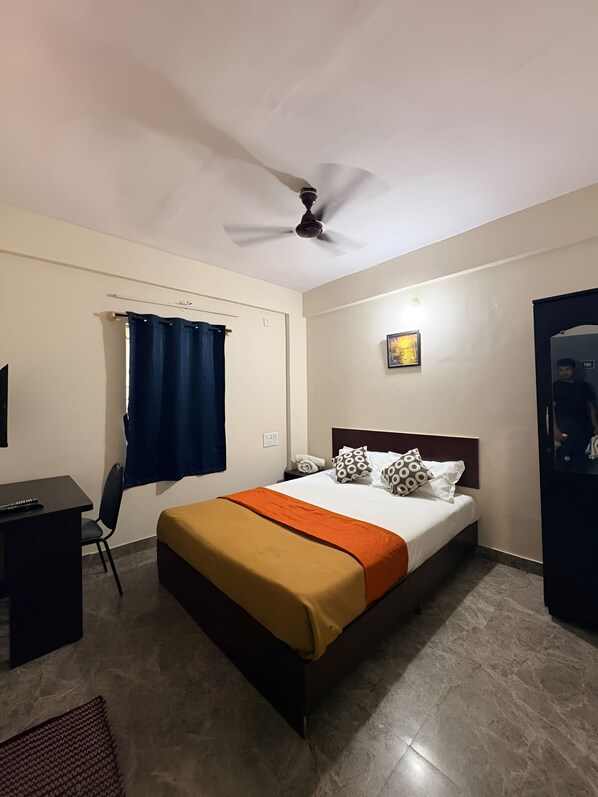 A hotel room with a bed, a desk, a chair, a ceiling fan, and a window with curtains. - MALABAR INN (Bengaluru)