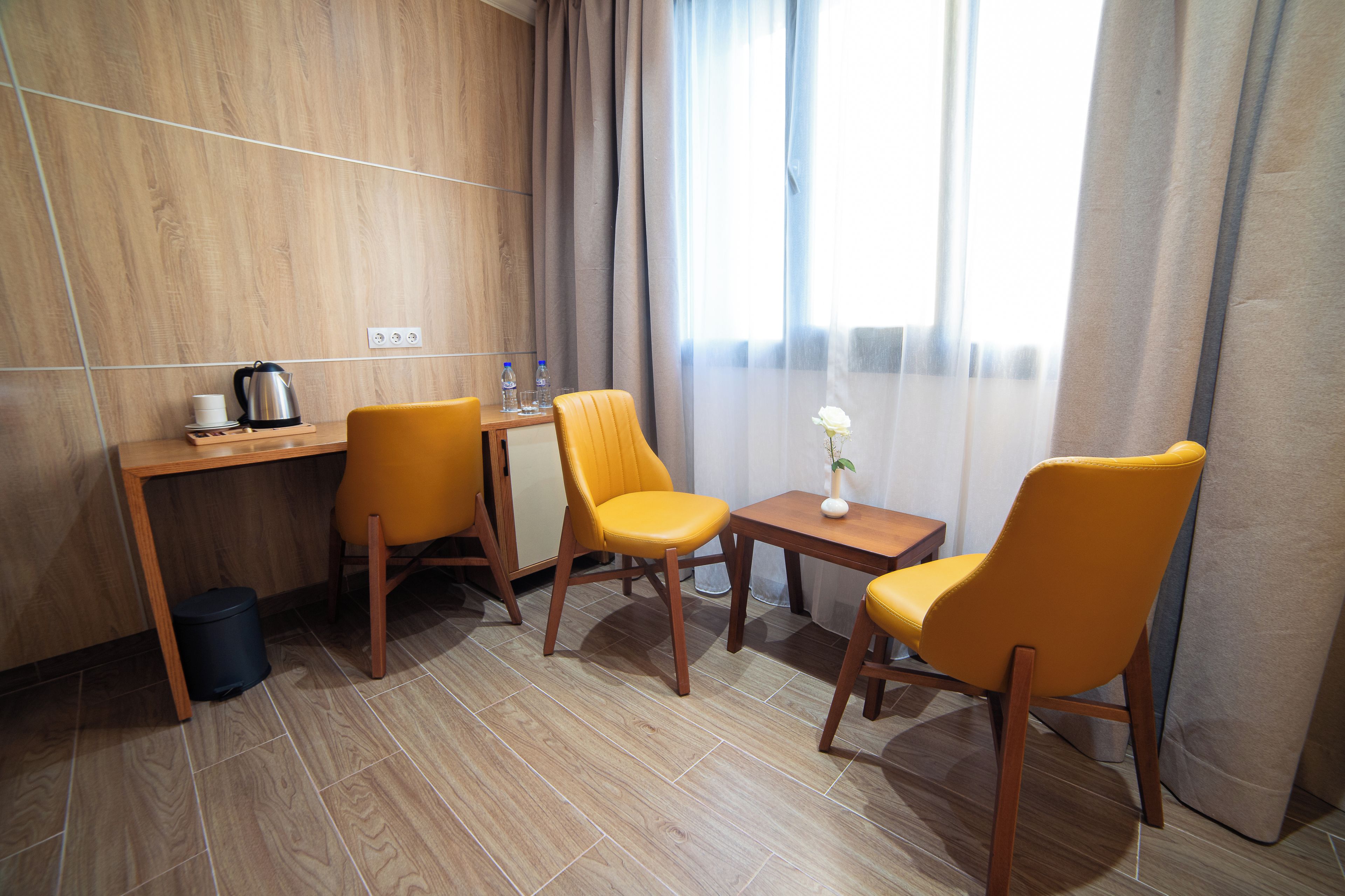 Una habitación con un escritorio de madera, dos sillas amarillas, una mesita con un florero y una ventana con cortinas.