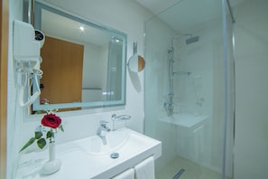 A bathroom with a white sink, a mirror, a hairdryer, a shower, and a vase with a red rose. - Barkat hôtel (Algiers)