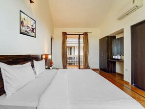 A hotel room with a large bed, a nightstand, a painting, a balcony with curtains, and a wardrobe. - Classic Aveeno Villas (Saligao)