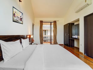 A hotel room with a large bed, a nightstand, a painting, a balcony with curtains, and a wardrobe. - Deluxe Aveeno Villas (Saligao)