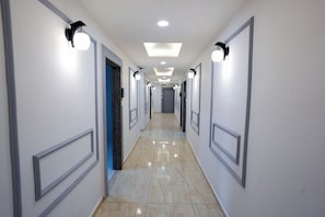 Um corredor comprido e estreito com paredes brancas, molduras de porta cinzas e chão de azulejos brilhantes.