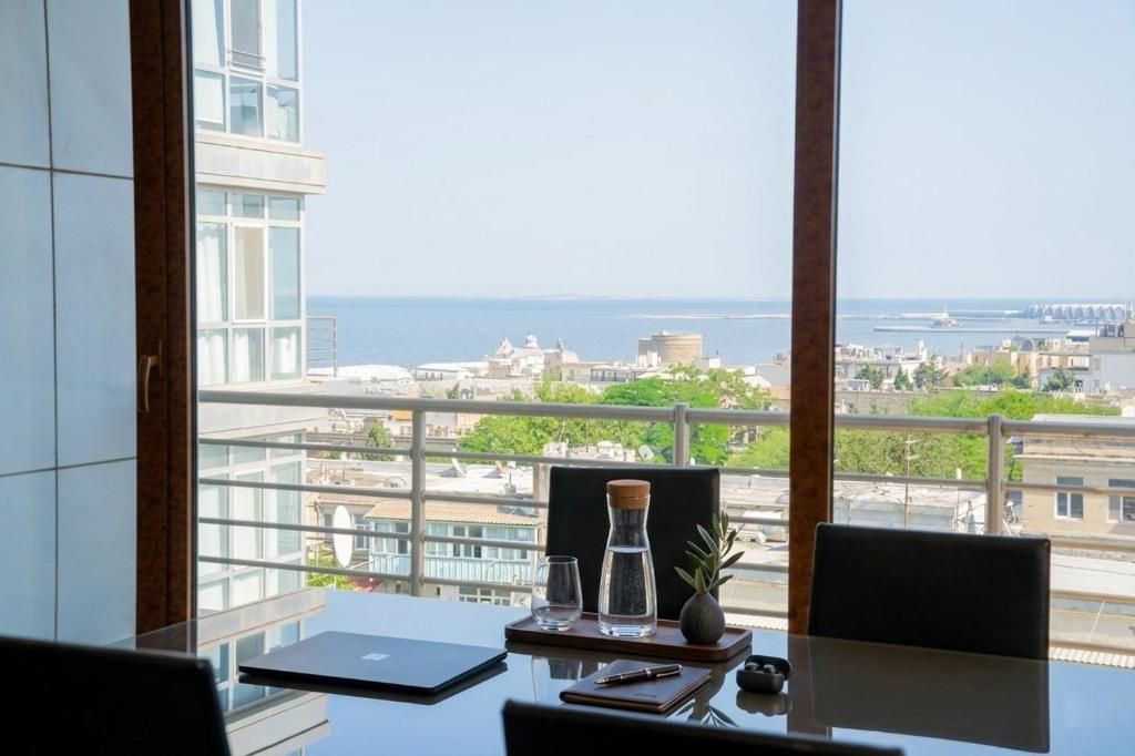 Un salón de conferencias con vistas al mar y al paisaje urbano, una computadora portátil, un vaso de agua y un cuaderno sobre la mesa.