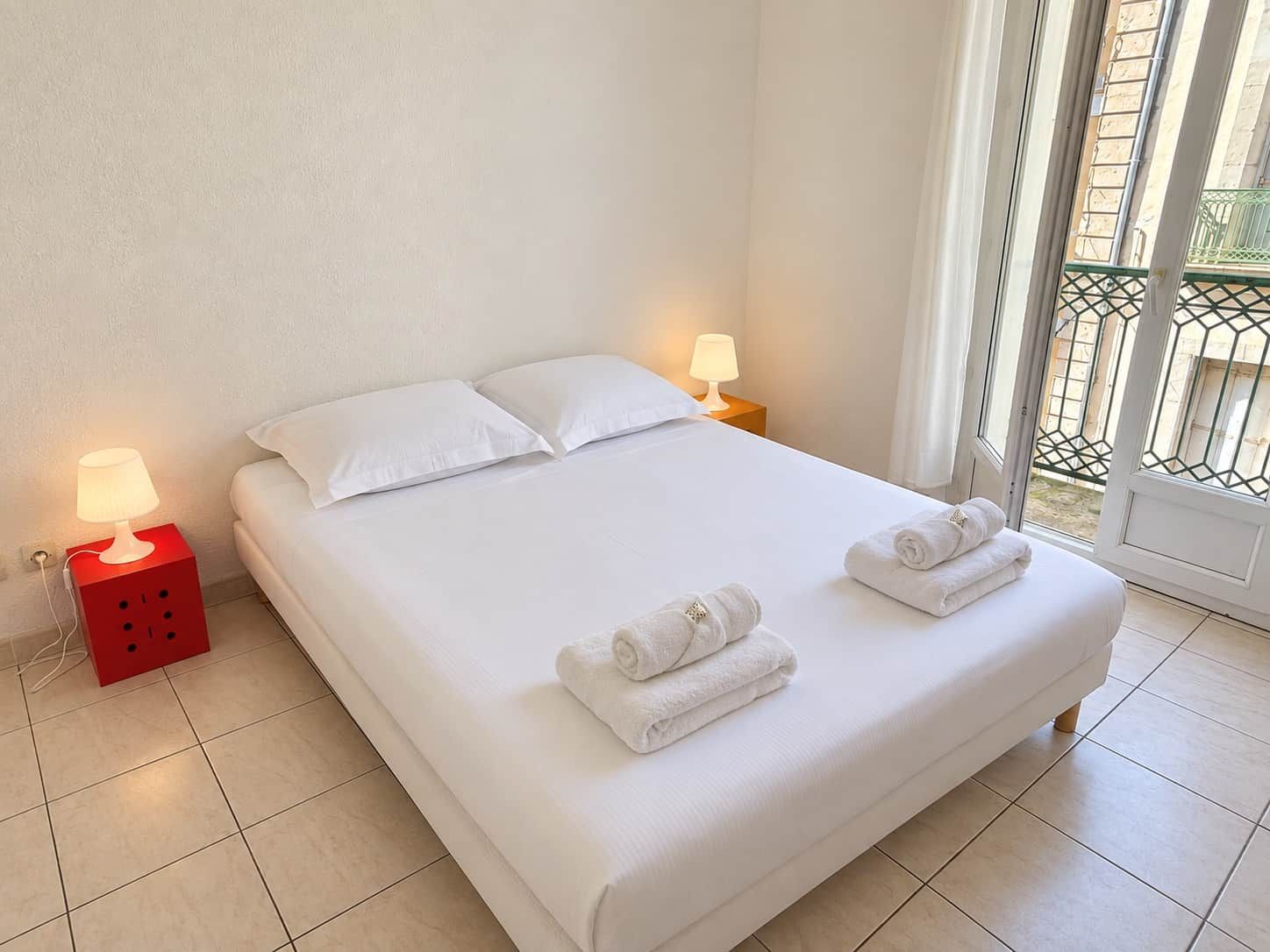A neatly made bed with white linens, two pillows, and two rolled towels on the bed. There are two bedside lamps on red nightstands, and a window with a view of a balcony.