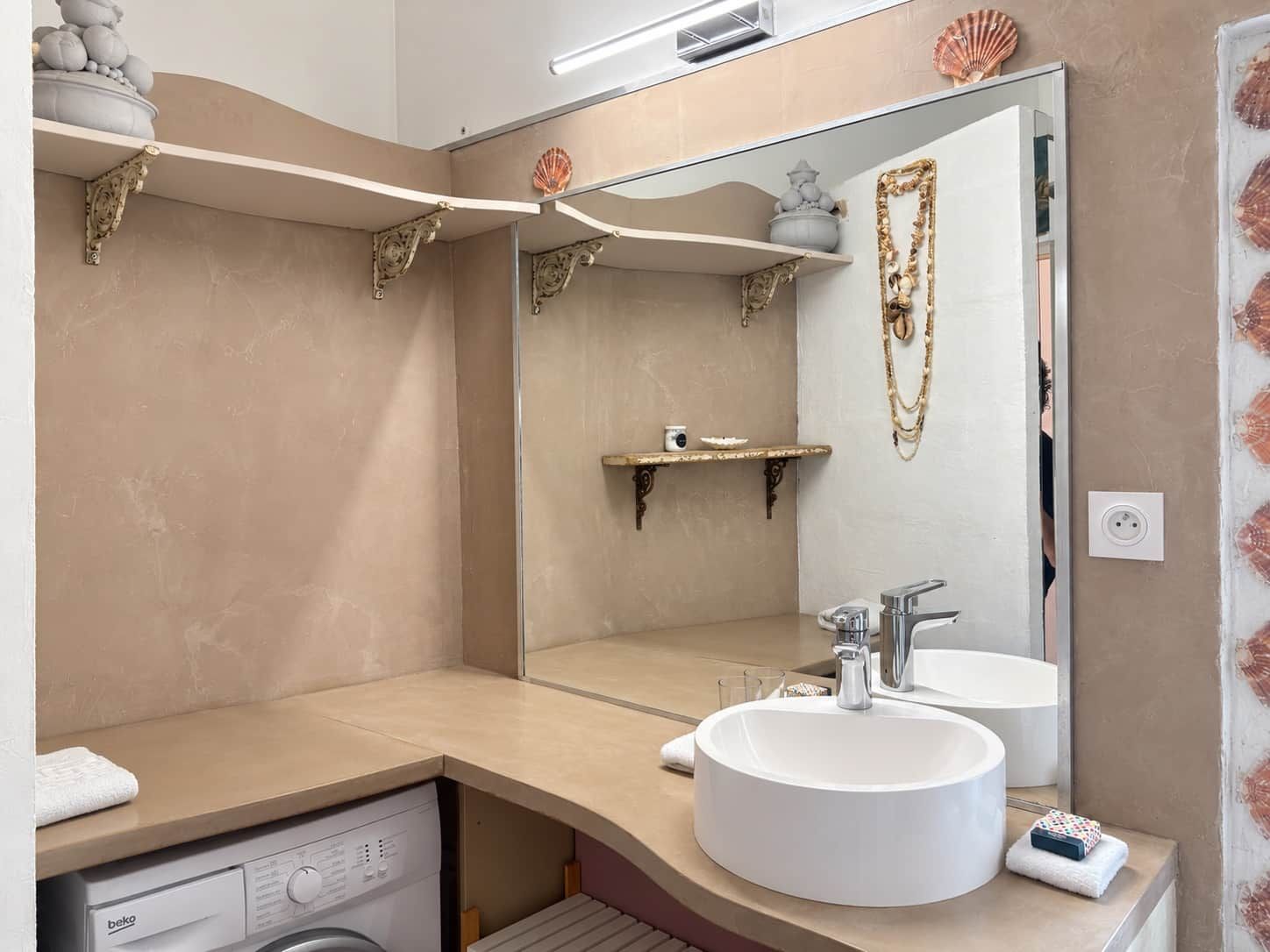 Een badkamer met een grote spiegel, een rond wastafel, een wasmachine en decoratieve schelpen aan de muur.