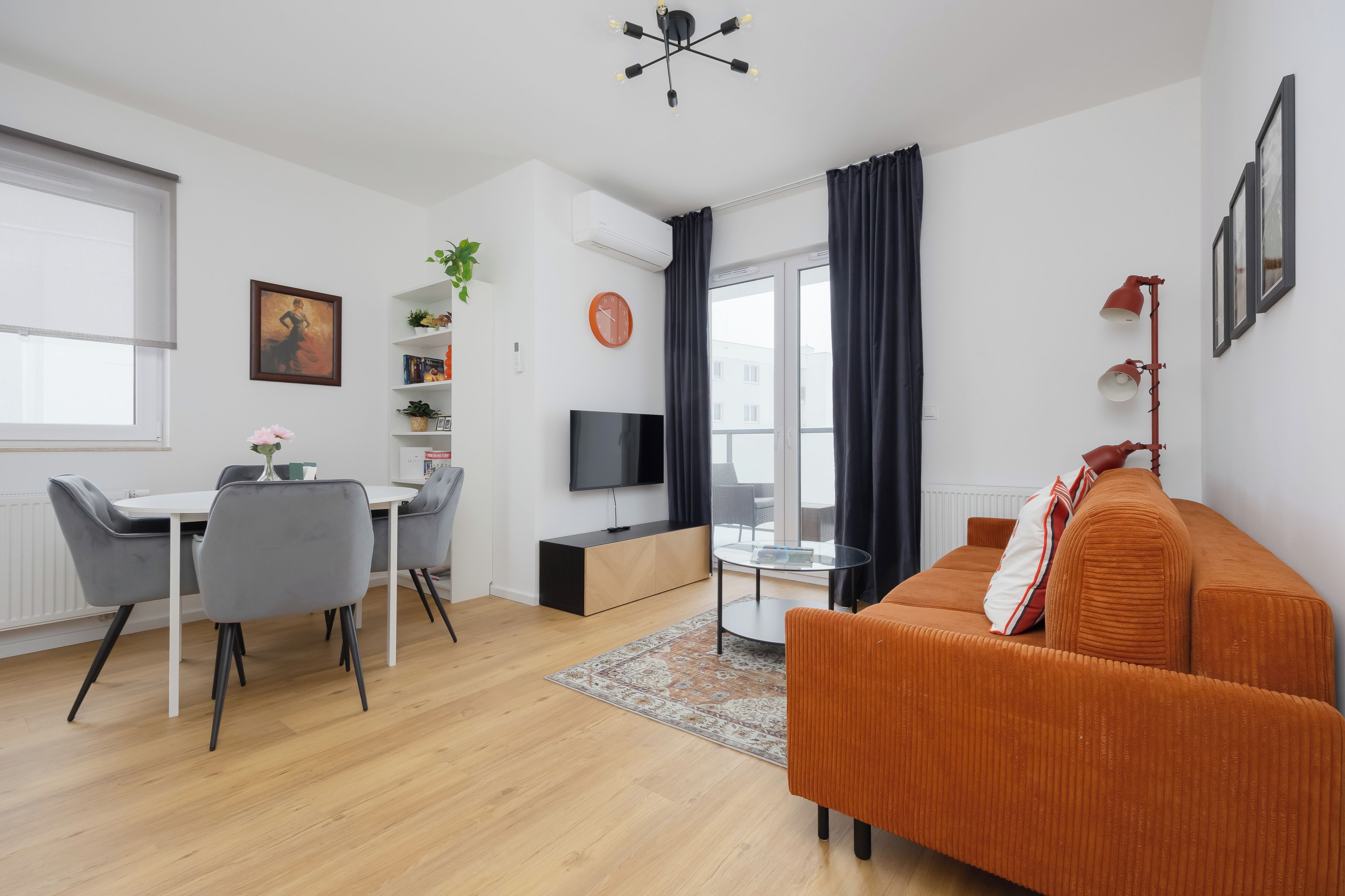 Ein modernes Wohnzimmer mit einer Couch, einem Esstisch mit Stühlen, einem Fernseher und einem großen Fenster mit Vorhängen.