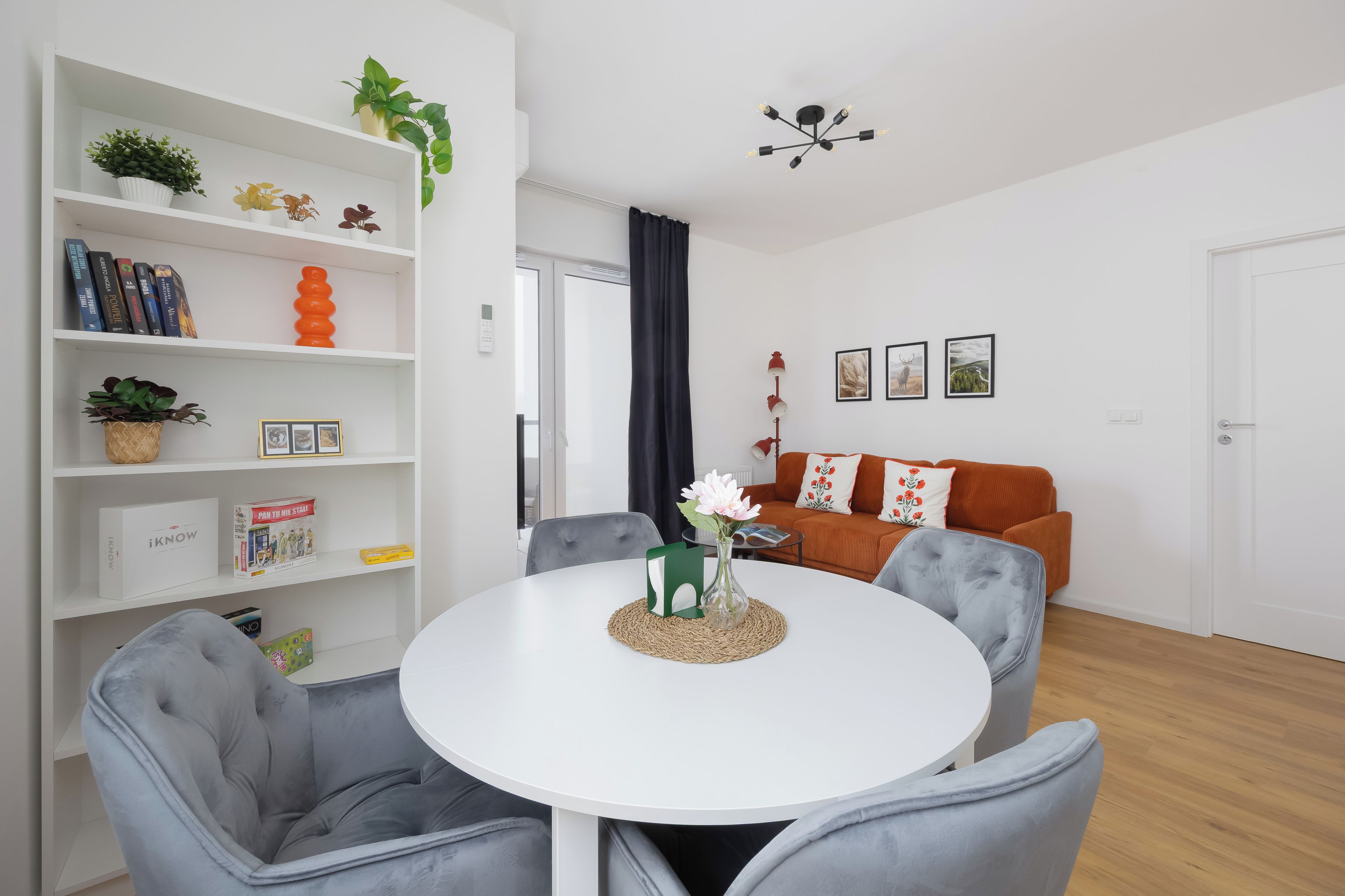 Ein modernes Wohnzimmer mit einem weißen runden Tisch, grauen Sesseln, einer Bücherwand mit Büchern und Dekoration, einer roten Stehlampe und einem braunen Sofa mit floralen Kissen.