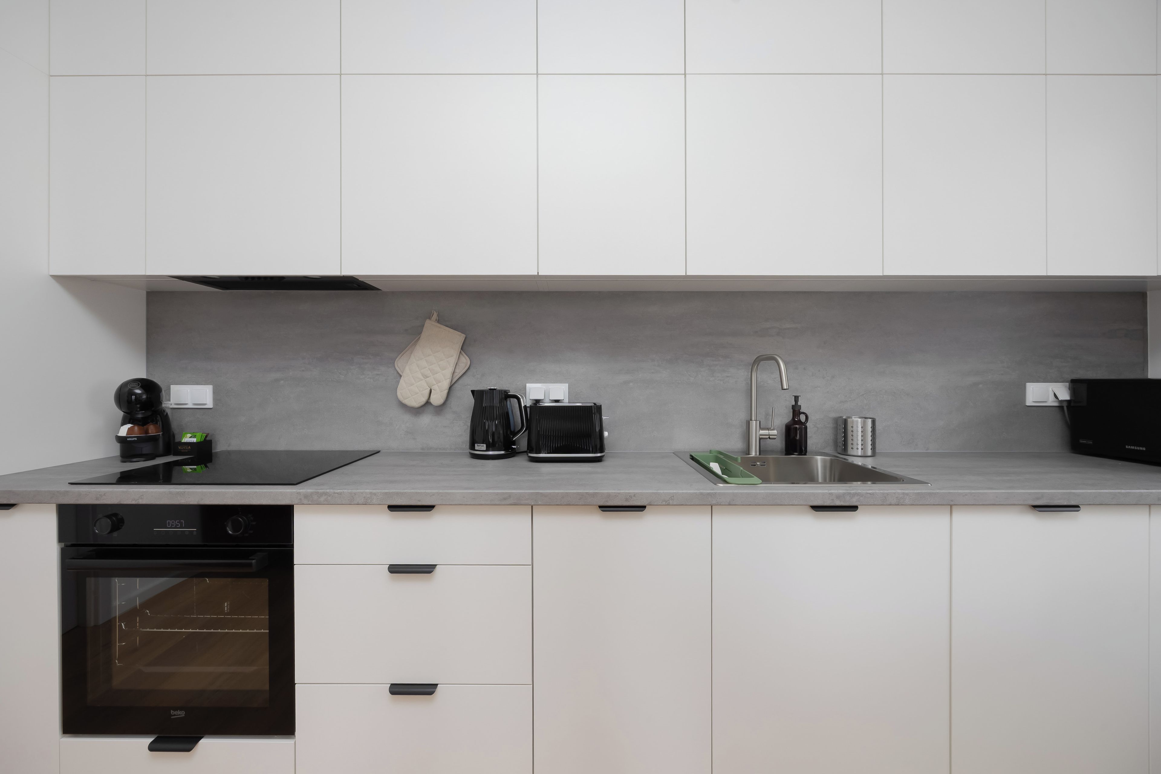 A modern kitchen with white cabinets, a built-in oven, a coffee maker, a kettle, and a sink.