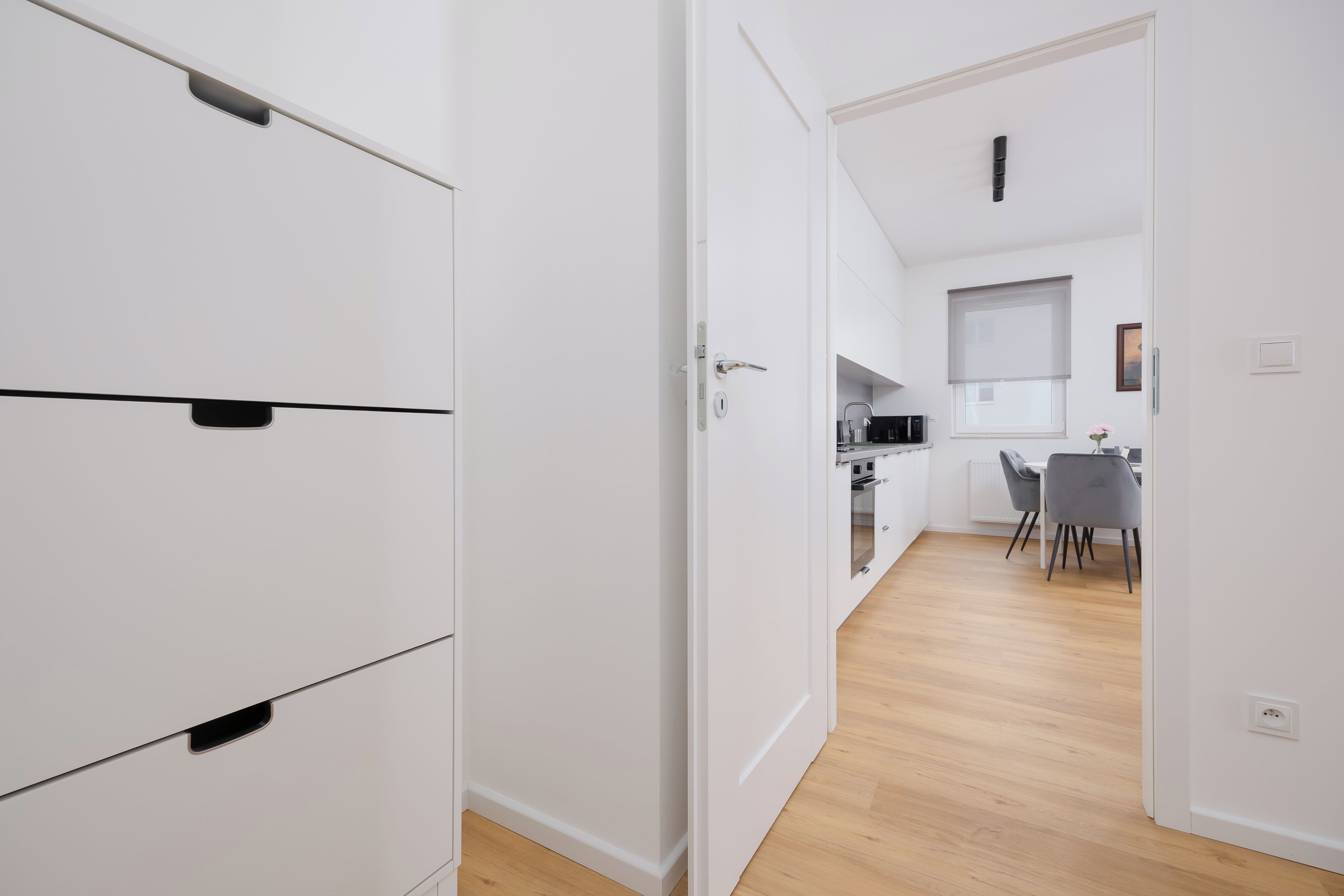 A modern kitchen with white cabinets, a dining area with a table and chairs, and a hallway leading to a bedroom.