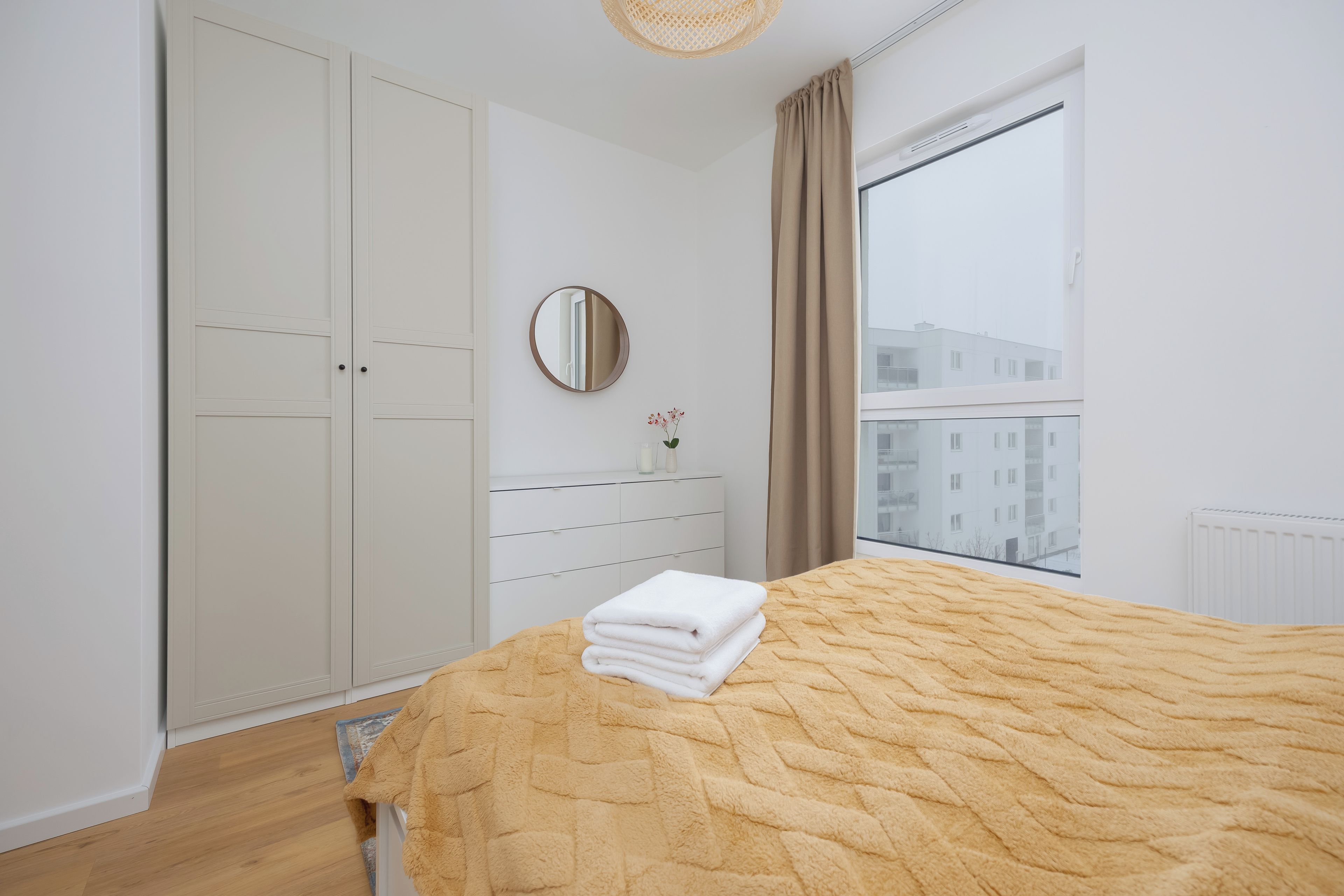 A bedroom with a bed, a wardrobe, a mirror, a dresser, and a window with a view of buildings.