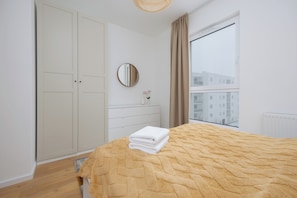 A bedroom with a bed, a wardrobe, a mirror, a dresser, and a window with a view of buildings.