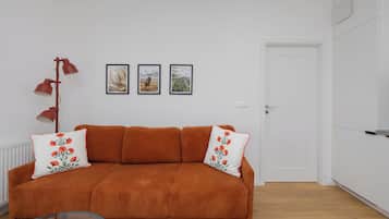 A room with a brown sofa, two white pillows with red floral designs, a red floor lamp, and two framed pictures on the wall.