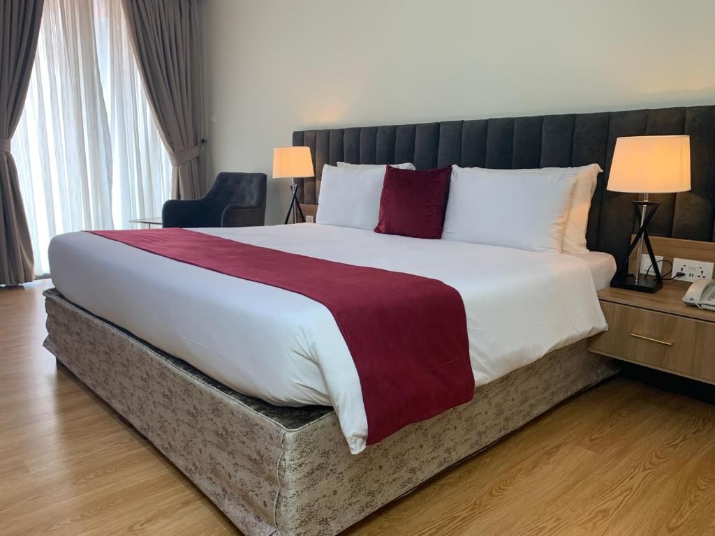 Una habitación de hotel con una cama grande, dos lámparas de noche, un cabecero y una ventana con cortinas.