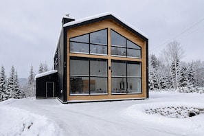 Eine moderne, zweistöckige Holzhütte mit großen Fenstern und einer Holzfassade, umgeben von schneebedeckten Bäumen.