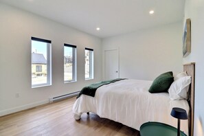 Quarto com cama, mesa de cabeceira, abajur e três janelas.