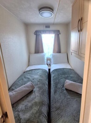 A narrow room with two beds, a window with curtains, a ceiling light, and a wardrobe.