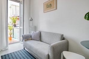 A room with a sofa, a small table, a radiator, a window with a view of buildings, and a potted plant. - Hostmate- Milano-Minimal Nest (Milan)
