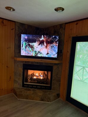 A living room with a stone fireplace, a flat-screen TV mounted above it, and a window with blinds. - Cozy Mountain Cabin Retreat in Murphy, NC (Murphy)