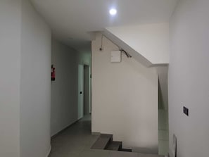 Un pasillo con una escalera que lleva a una plataforma, un detector de incendios a la izquierda y un accesorio de iluminación en el techo.