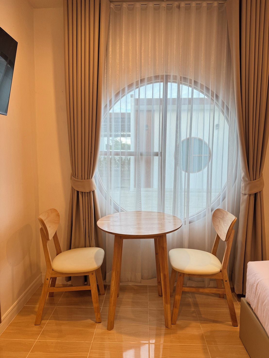 Una habitación con una mesa redonda de madera, dos sillas, una ventana con cortinas y una cama.