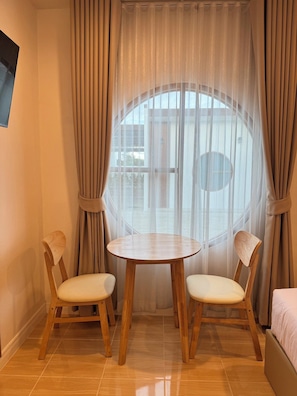 A room with a round wooden table and two chairs, a window with curtains, and a bed. - Feel at home resort  (Nakhon Pathom)