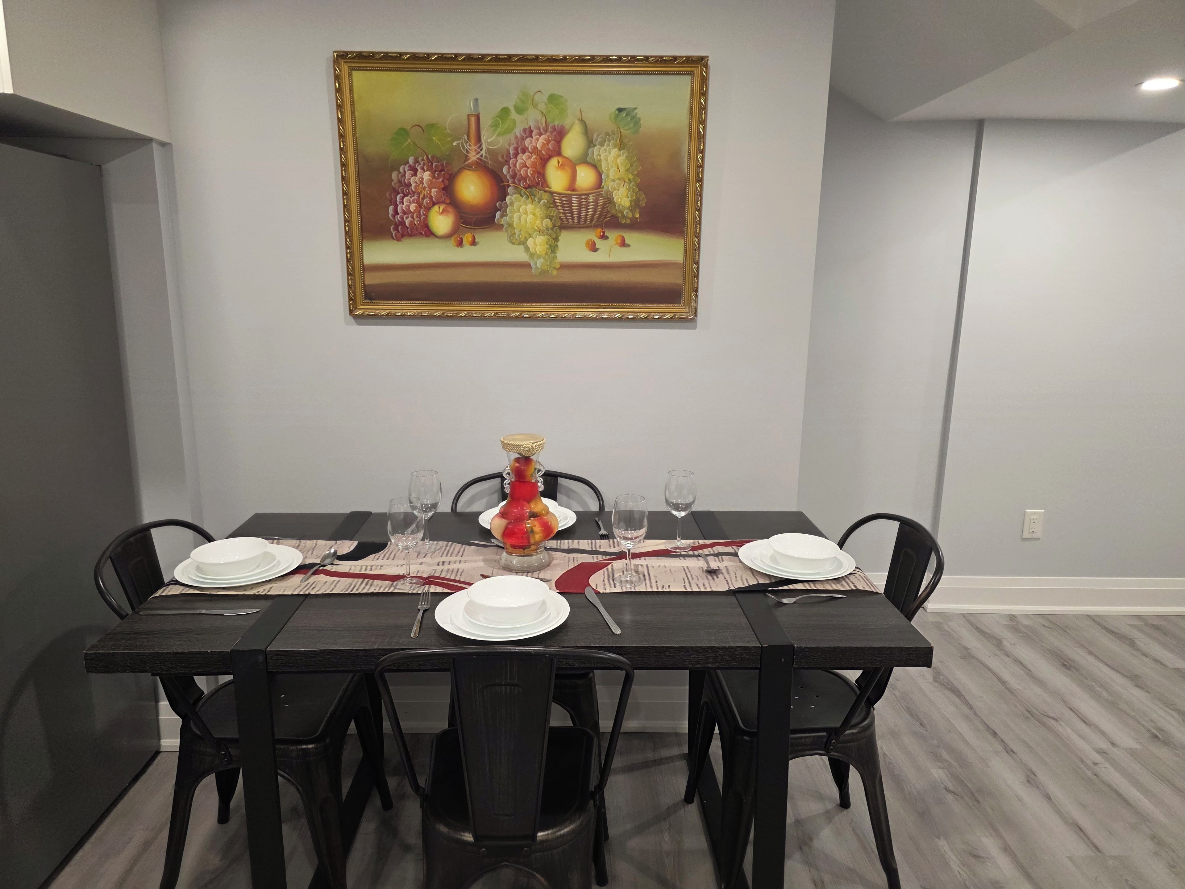 Un coin repas avec une table dressée pour quatre personnes, des chaises, un tableau au mur et un vase rempli de fruits.