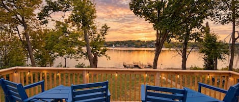 A deck with two blue chairs and a table, overlooking a lake with trees and a distant bridge.