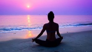 Eine Person sitzt in einer Meditationshaltung an einem Strand, im Hintergrund das Meer und ein Sonnenuntergang.