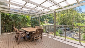 Una terraza de madera con una mesa y sillas, rodeada por una valla metálica y paneles de vidrio, con vistas a un jardín.
