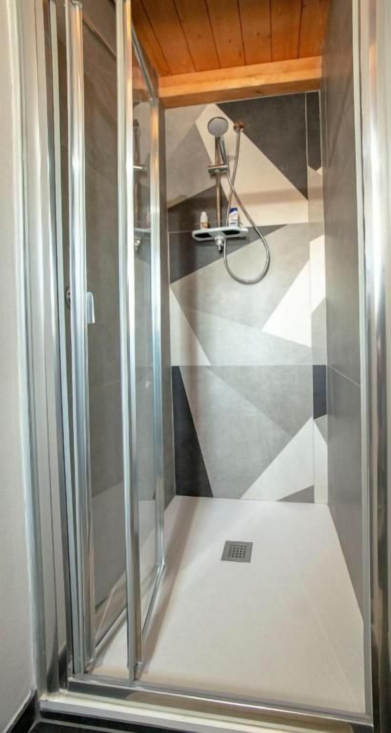 Box doccia in vetro con soffione e pannello comandi, pavimento piastrellato e soffitto in legno.