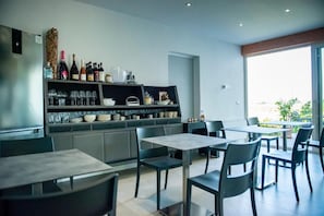 A modern dining area with tables and chairs, a kitchen with wine bottles and glasses, and a large window offering a view. - FORESTERIA LOMBARDA BED AND BIKE CREMONA (Crotta d'Adda)