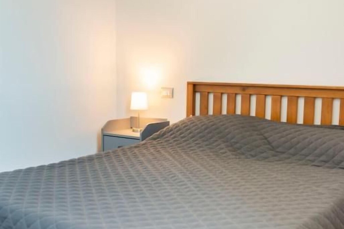 A bed with a wooden headboard, a nightstand with a lamp, and a plain wall.