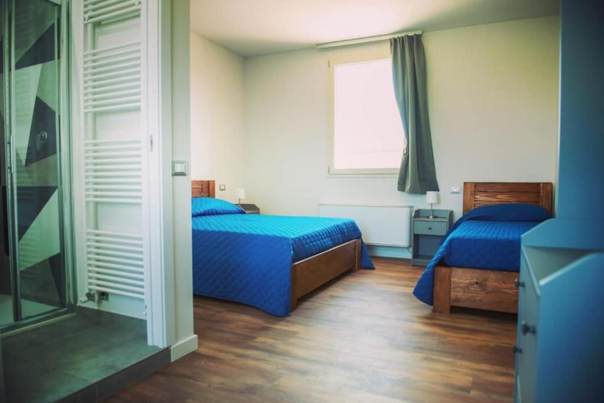 A bedroom with two beds, a window with curtains, a bedside table, and a radiator.