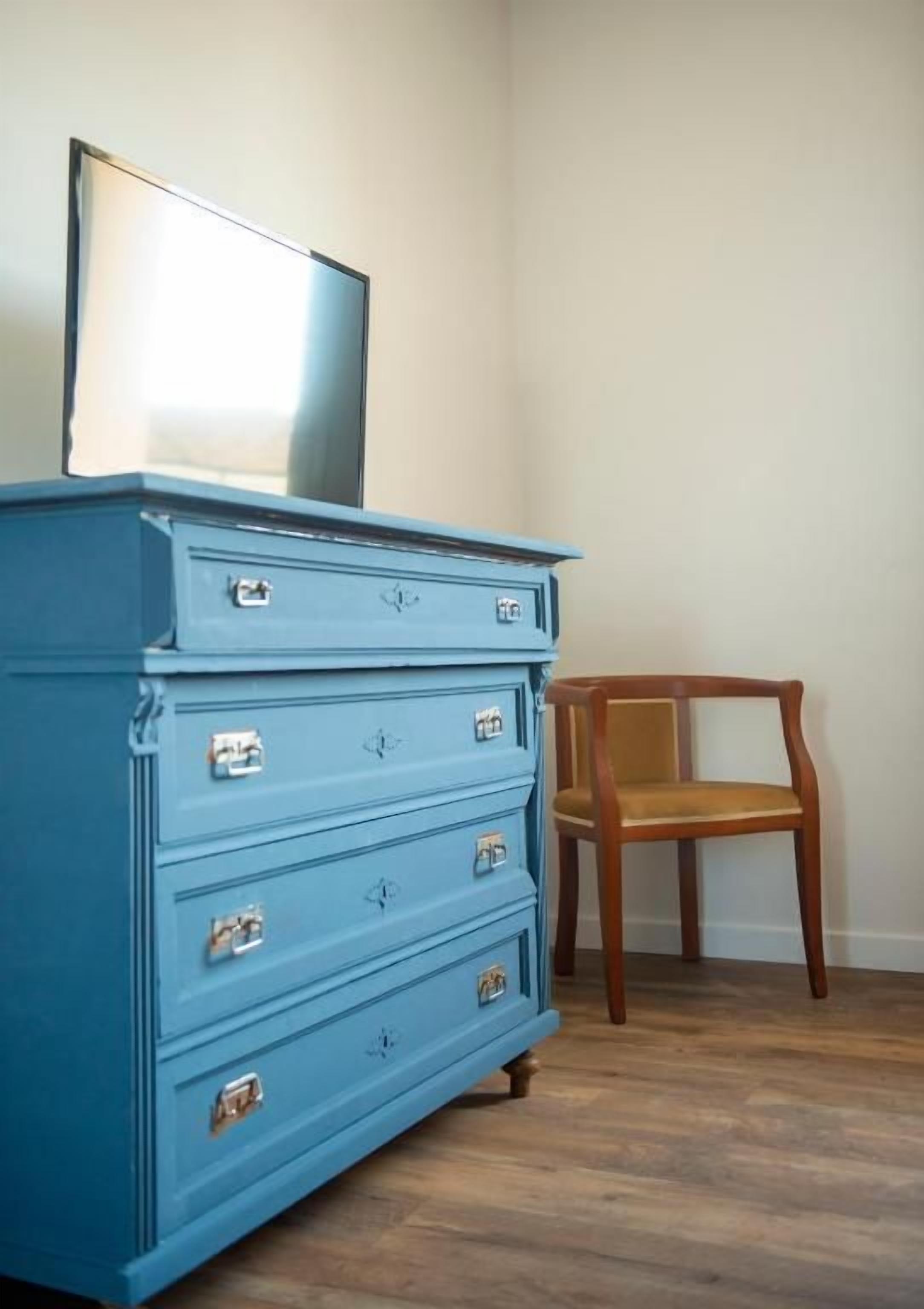A blue dresser with drawers and a mirror, a wooden chair, and a wooden floor.