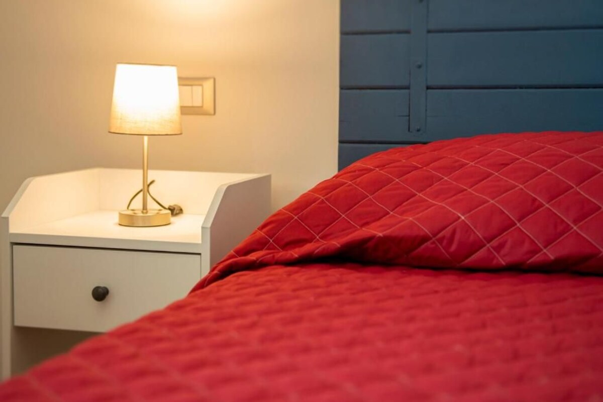 A bed with a red quilted cover, a white bedside table with a lamp, and a blue headboard.