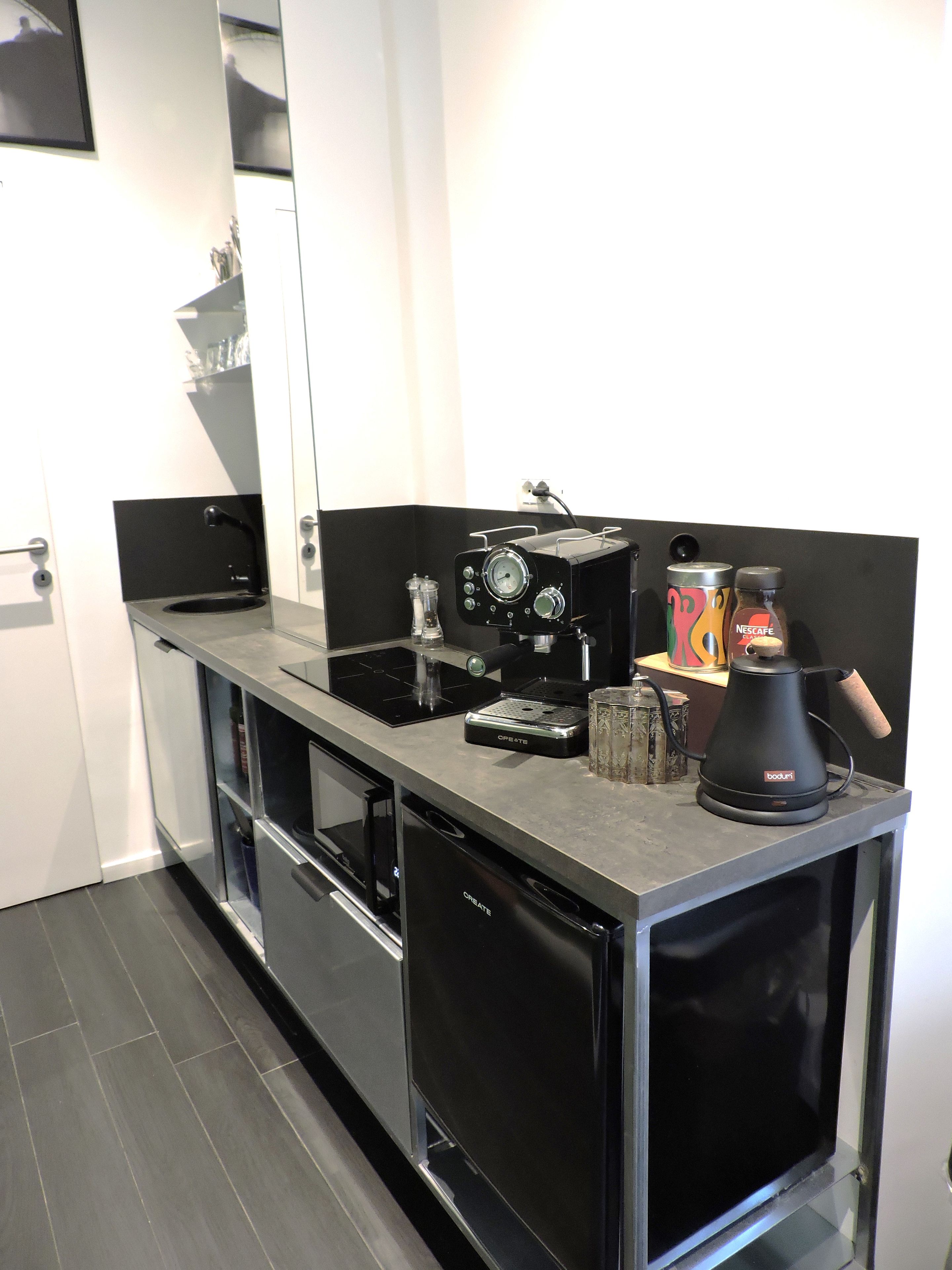 Une petite cuisine avec un plan de travail noir, une machine à café, un verre et une boîte de Nescafe.