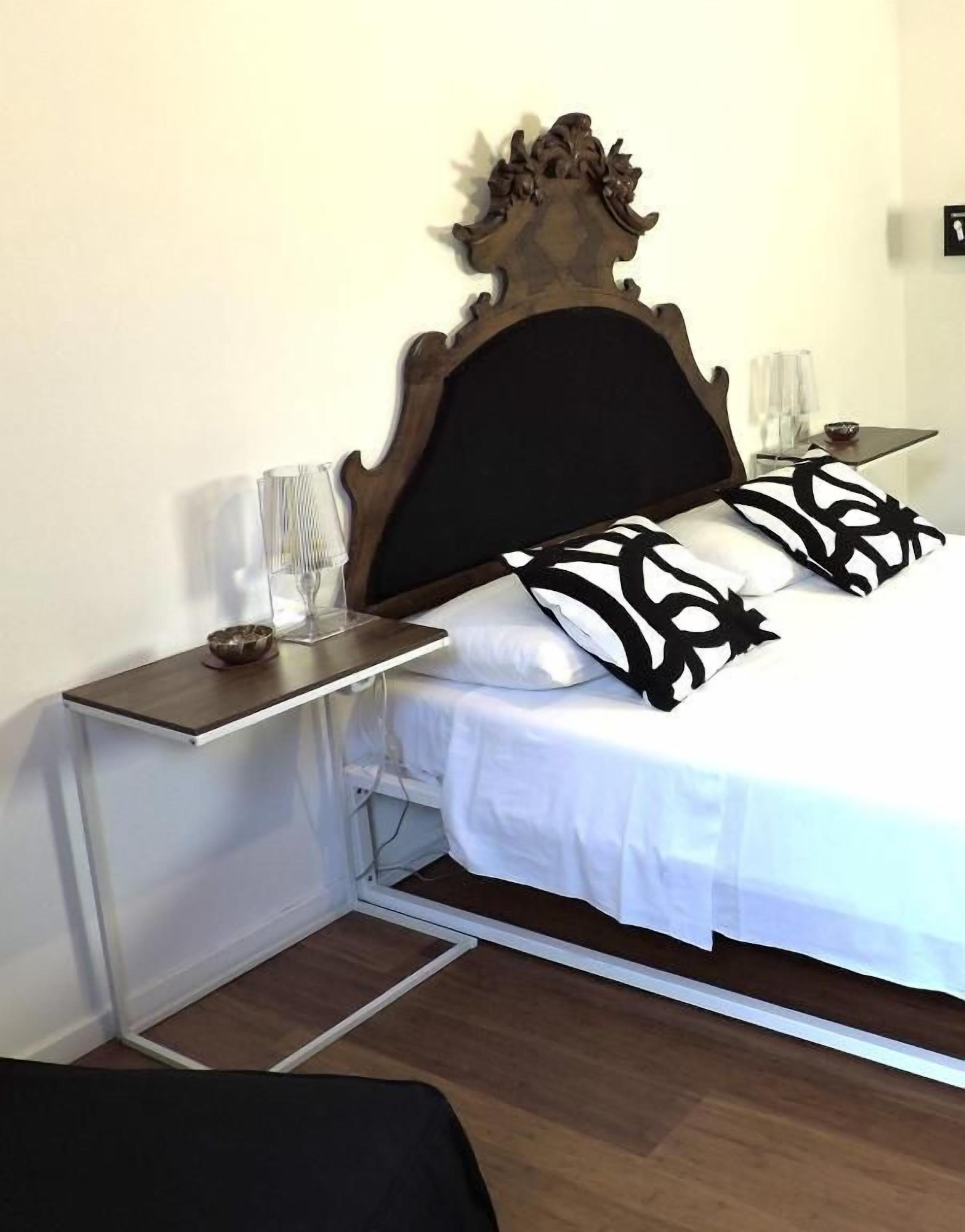 Una cama con cabecera de madera, ropa de cama blanca y almohadas negras estampadas. Hay una mesita de noche con una lámpara y un pequeño objeto decorativo.