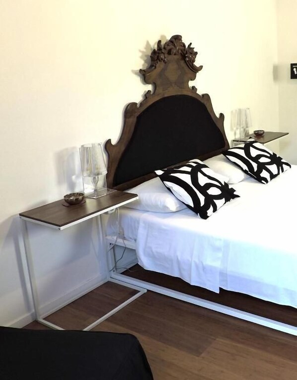 A bed with a wooden headboard, white bedding, and black patterned pillows. There is a bedside table with a lamp and a small decorative item. - Art Lab Residence (ROMA)