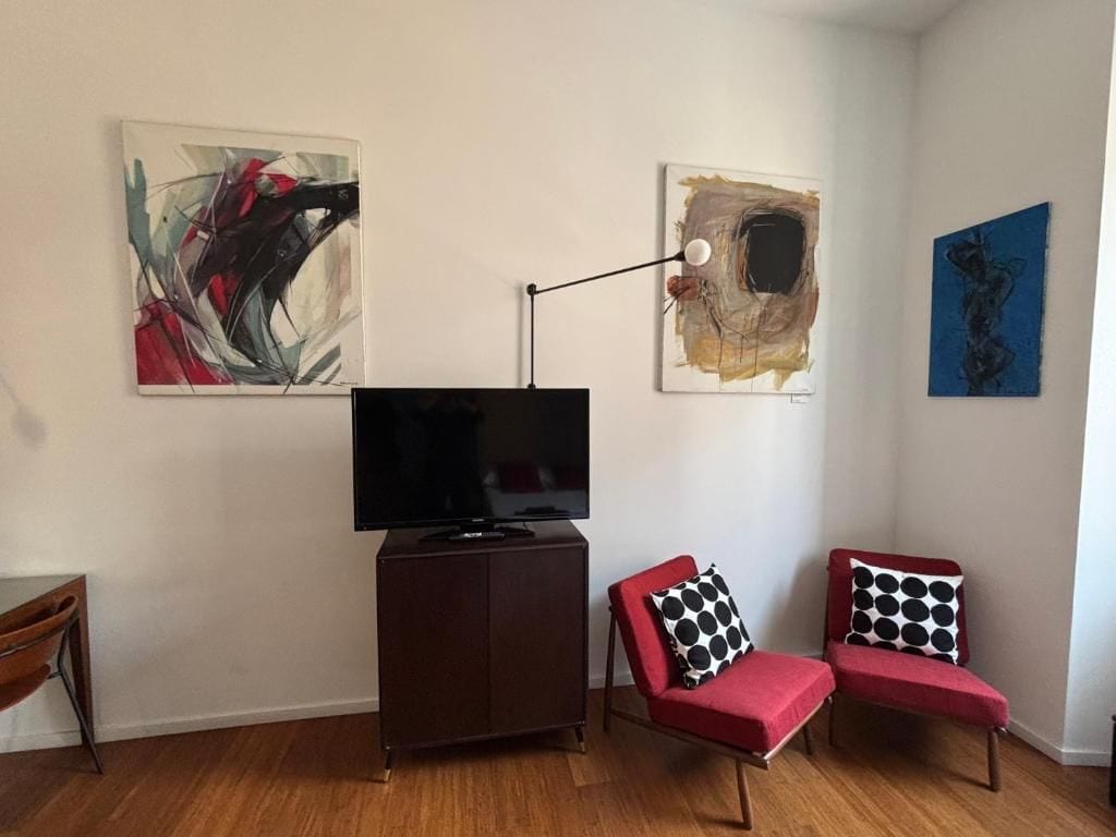 Una sala de estar con un televisor de pantalla plana sobre un mueble bajo, dos sillones rojos con cojines de estampado blanco y negro, y arte abstracto en las paredes.