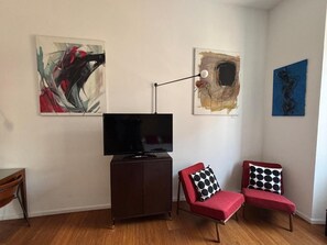 Ein Wohnzimmer mit einem Flachbildfernseher auf einem Schrank, zwei rote Sessel mit Kissen in Schwarzweißmuster und abstrakten Kunstwerken an den Wänden.