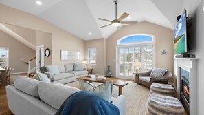 A spacious living room with a high ceiling, a ceiling fan, a fireplace, and a large window with a view. - Beautiful 5 Bedroom Home with a pool in Holgate! (Beach Haven)