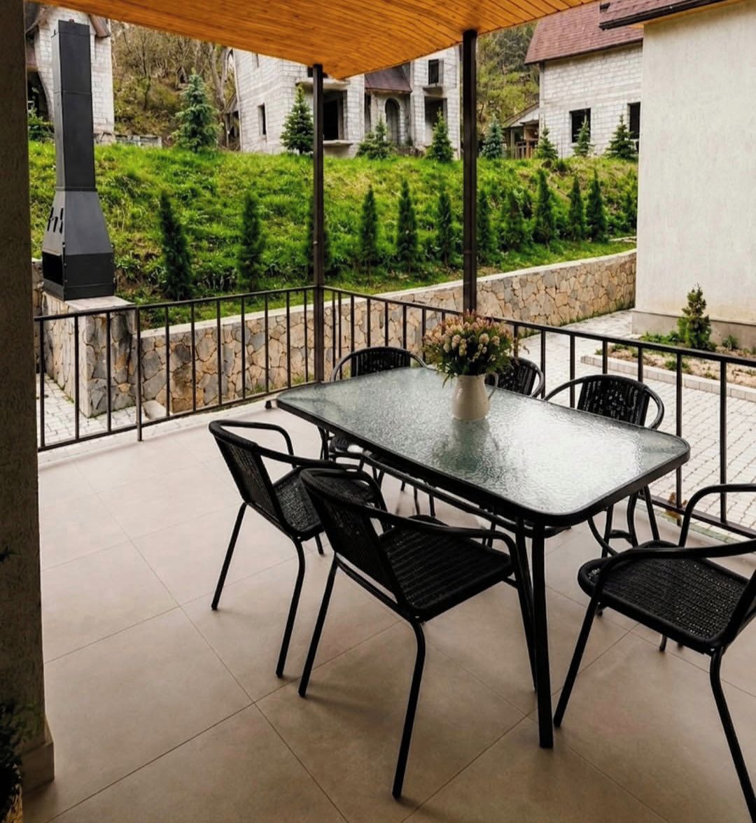 Un patio esterno coperto con tavolo e sedie in vetro, un vaso di fiori e vista su un edificio in pietra e vegetazione.