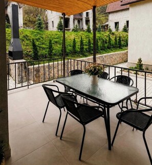 Un patio techado al aire libre con mesa y sillas de vidrio, un florero y vistas a un edificio de piedra y follaje.