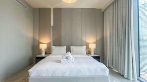 Ein Hotelzimmer mit einem großen Bett, zwei Nachttischen mit Lampen, einem gefalteten Handtuch auf dem Bett und Blick auf ein Gebäude durch das Fenster.