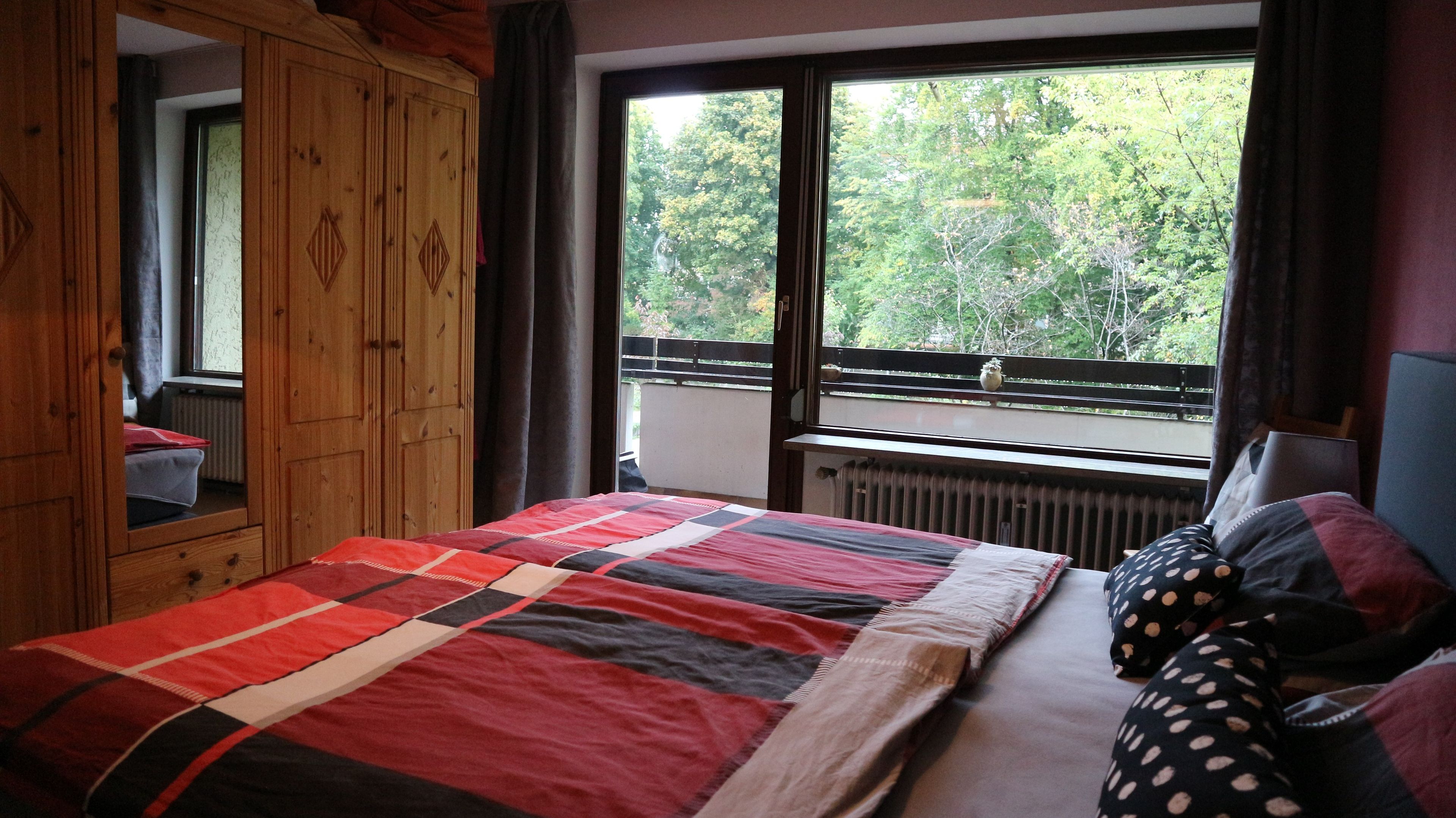 Un dormitorio con cama, un armario de madera, un ventanal con vistas a árboles y un radiador.