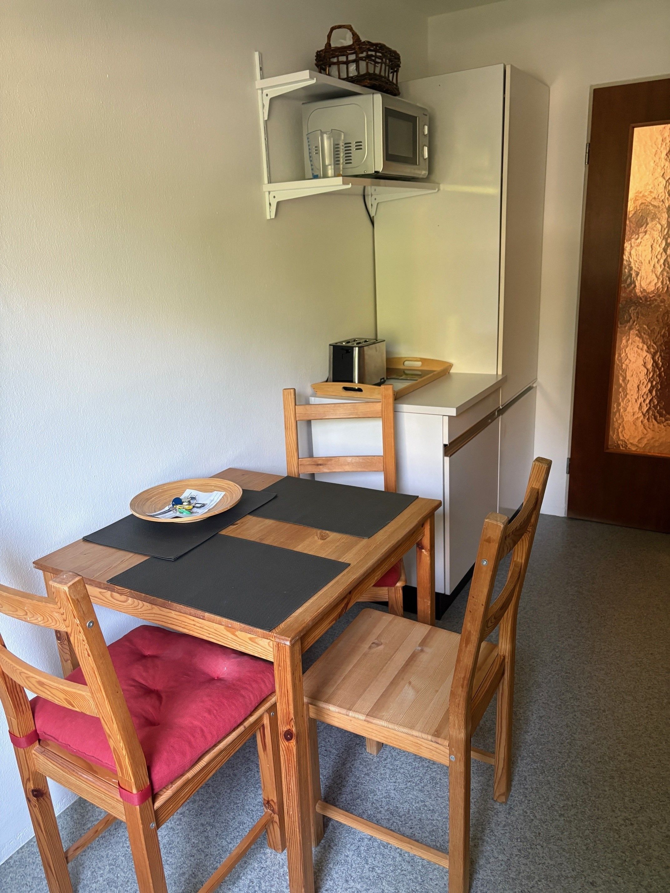 Una kitchenette compacta con microondas, tostadora y una pequeña encimera. Hay una mesa de comedor de madera con sillas, y un cojín rojo en una de ellas.
