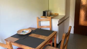 Una kitchenette compacta con microondas, tostadora y una pequeña encimera. Hay una mesa de comedor de madera con sillas, y un cojín rojo en una de ellas.